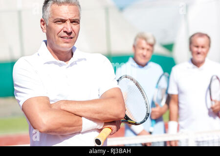 Portrait of confident mature man holding tennis racket while standing with arms crossed against friends on court during sunny day Stock Photo