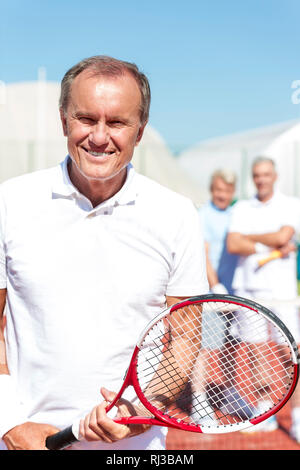 Portrait of smiling senior man holding tennis racket while standing against mature friends on court during sunny day Stock Photo