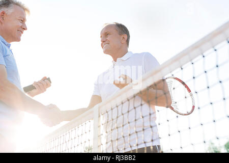 Low angle view of smiling men shaking hands while standing at tennis court against clear sky Stock Photo