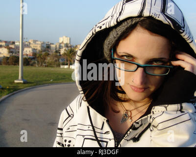 Young woman with blue green eyes and glasses, looking at the camera with a serious expression, in the forefront of a city park with building complexes Stock Photo