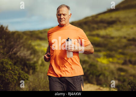 fitness man jogging outdoors holding a water bottle. Portrait of a senior man running outdoors with a hill in the background. Stock Photo