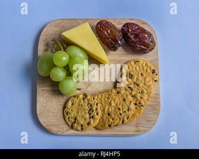 Seeded crackers, cheese, grapes, and dates, arranged on a wooden board with a blue background. Stock Photo