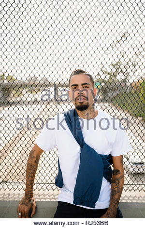 Portrait confident, tough Latinx young man with tattoos leaning against overpass fence