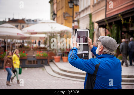 Tourist taking photo with iPad, old bearded man in blue jacket and grey beret photograph Royal Castle Square full of tourists, tourism season in Plac  Stock Photo