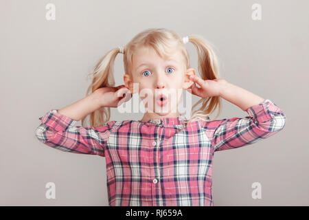 Closeup portrait of funny blonde Caucasian preschool girl making faces in front of camera. Child holding pulling dragging ears on light background. Ki Stock Photo