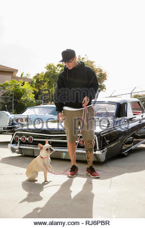Latinx young man with dog in front of low rider car in sunny parking lot