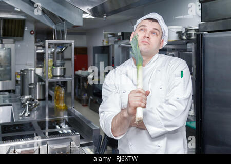 Closeup man professional chef in white uniform composing a recipe, thinks what to cook and holds the stem of leek in his hand. Quality control concept Stock Photo