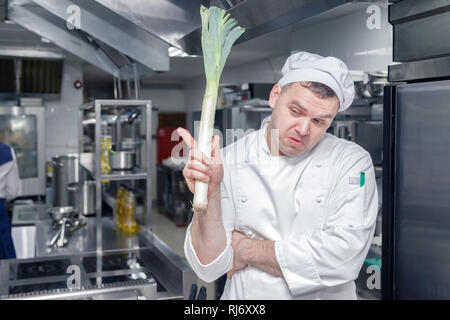 Closeup man professional chef in white uniform composing a recipe, thinks what to cook and holds the stem of leek in his hand. Quality control concept Stock Photo