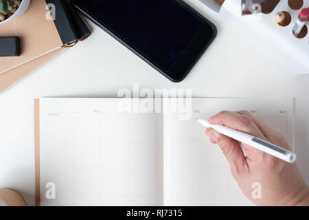 top view of hand writing on open calendar planner for business resolution with modern office stationery and take away coffee cup on white desk in offi Stock Photo
