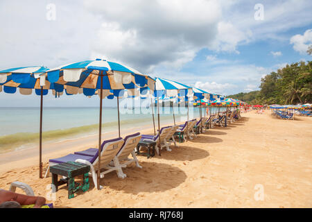 Row of sun loungers and colourful blue and white sunshades and parasols on the sandy shoreline at Surin Beach, west coast of Phuket, Thailand Stock Photo