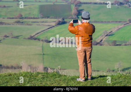 Lone man holding compact camera up taking photos of a landscape view of fields in England, UK. Stock Photo