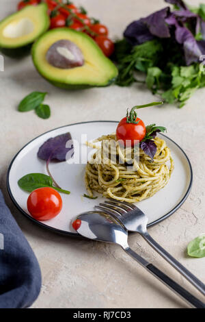 Tasty Italian pasta with pesto, herbs and cherry tomatoes at white plate, with fork and spoon and fresh herbs at white background Stock Photo