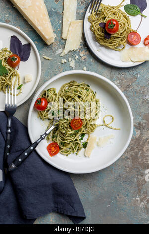 Italian spaghetti with pesto, herbs and cherry tomatoes at three plates at blue and gray background with forks, napkin and parmesan cheese Stock Photo