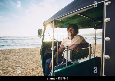 Young tuk tuk driver waiting for passenger against sand beach and sea in Sri Lanka. Stock Photo