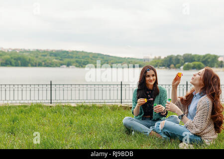 Two sisters having fun while making bubbles and sitting on the grass near the lake with a beautiful landscape in the background Stock Photo