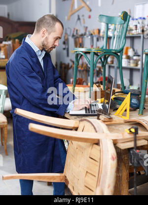 Furniture restorer using laptop in process of renovation vintage chair in workshop Stock Photo