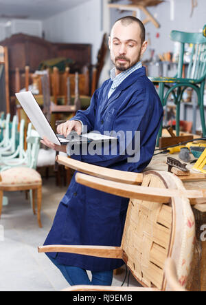 Furniture restorer using laptop in process of renovation vintage chair in workshop Stock Photo