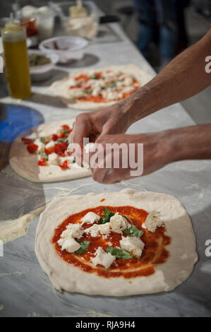 Preparing Pizza Margherita on a marble countertops. Pizzaiolo puts pieces of mozzarella and basil leaves over a raw pizza dough with tomato sauce. Stock Photo