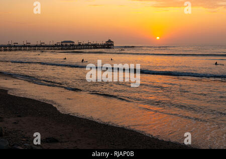 The wooden pier of Huanchaco beach near Trujillo with the silhouette of surfers at sunset, Peru. Stock Photo