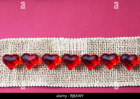 Dark pink valentine, wedding card background with a row of shiny hearts on a burlap string with tiny pearls Stock Photo