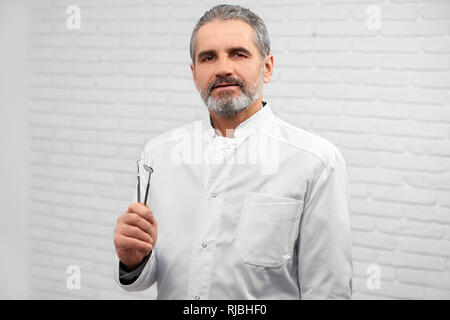Handsome man wearing in white medical uniform looking at camera. Mature stomatologist holding restoration instruments. Dentist posing in studio with white background. Stock Photo