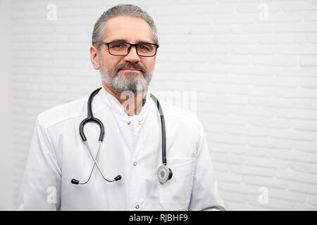 Professional ENT doctor looking at camera and posing in studio with white background. Mature bearded physician wearing in white medical uniform and glasses, having stethoscope on neck. Stock Photo