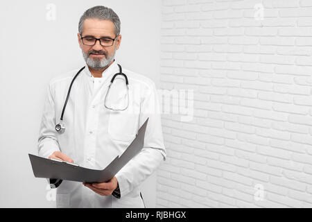 Professional ENT doctor standing in studio with white background. Mature, bearded doctor holding folder, looking down and reading. Physician having stethoscope on neck. Stock Photo