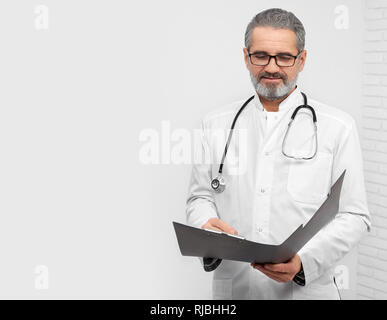 Confident ENT doctor holding folder, looking down and reading. Professional therapist with stethoscope on neck wearing in medical white coat and glasses. Studio with white background. Stock Photo