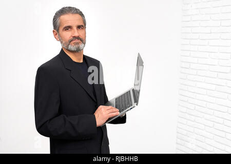 Bearded man holding modern notebook in hands, looking at camera. Mature, serious man wearing in stylish black outfit. Businessman posing in studio with white background. Stock Photo