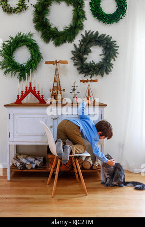 Boy kneeling on a chair in front of a sideboard stroking his cat Stock Photo