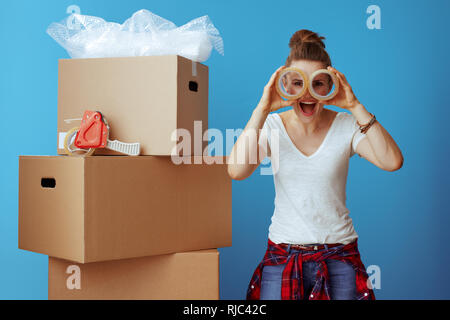 happy modern woman in white t-shirt near cardboard box looking through adhesive tapes isolated on blue Stock Photo