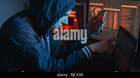 The male hacker in the hood is pointing his finger at the display indicating the location of the cyberattack and hacking data on the screen background Stock Photo