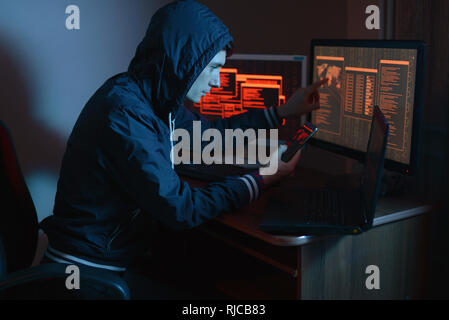Male hacker in the hood holding the phone in his hands trying to hack the mobile device cloud and steal data in the dark under neon light. The concept Stock Photo