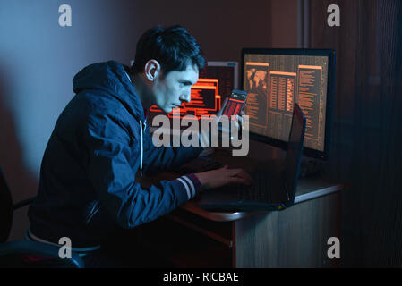 Male hacker holding the phone in his hands trying to hack the mobile device cloud and steal data in the dark under neon light. The concept of cyber se Stock Photo
