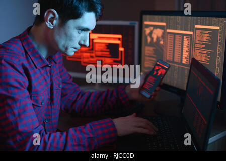Hacker holding the phone in his hands trying to hack the mobile device cloud and steal data in the dark under neon light. The concept of cyber securit Stock Photo