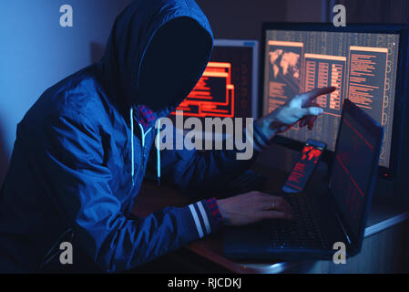 An anonymous hacker with no face typing the code tries to hack the system and steal accesses on the background screens in the dark. The concept of cyb Stock Photo