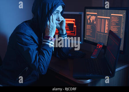 A male hacker in a hood thinking about the problem of hacking or malware infection on the background of screens with codes in neon light. The concept  Stock Photo