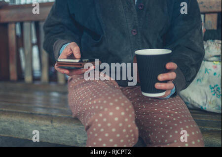 A young woman is sitting on a bench outdoors drinking take away coffee and using smartphone Stock Photo