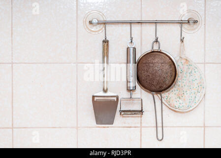 Kitchen utensils hanging on wall with sieve, metal peeler and towel mounted with suction cups closeup of tiled wall Stock Photo