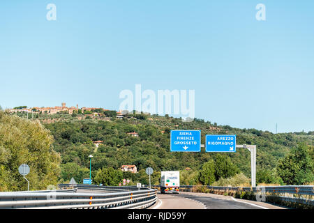 Seina, Italy - August 27, 2018: Hilltop town village cityscape in Tuscany during sunny summer day and highway road street with blue exit sign Stock Photo