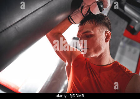 Tired young man in sports clothing training hard on heavy punch bag. Muscular sportsman with boxing gloves looking away while standing in boxing gym Stock Photo