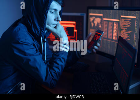 Hacker in a hood thinking about the problem of hacking or malware infection on the background of screens with codes in neon light. The concept of cybe Stock Photo