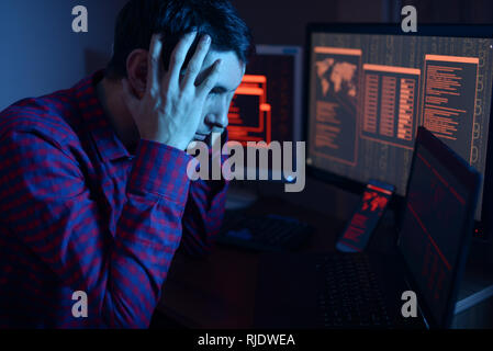 Male hacker thinking about the problem of hacking or malware infection on the background of screens with codes in neon light. The concept of cybersecu Stock Photo