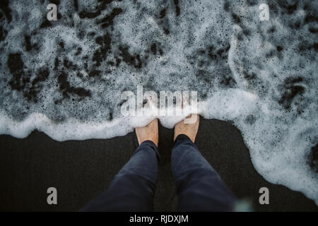 From above crop legs of anonymous traveler standing on wet sand near smooth sea foam Stock Photo