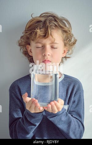 Sweet little boy keeping eyes closed and holding glass vase with transparent water while standing near white wall Stock Photo