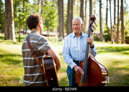 Smiling senior man playing a double bass while his grandson plays an acoustic guitar in a forest. Stock Photo
