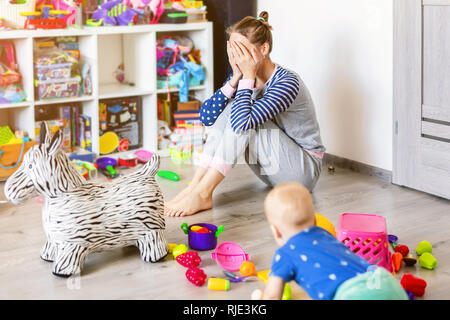 Tired of everyday household mother sitting on floor with hands on face. Kid playing in messy room. Scaterred toys and disorder. Happy parenting Stock Photo