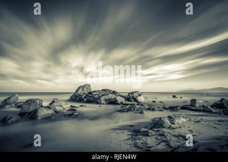 Long exposure black and white image of Mediterranean sea washing over rocks and onto sandy beach at sunrise near Ile Rousse in Corsica with orange glo Stock Photo