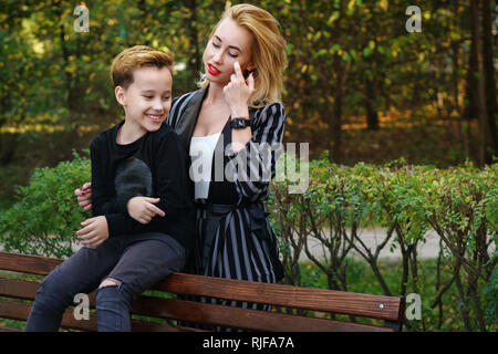 Young attractive mother and son in city park. Boy is sitting on bench and woman is hugging him from behind. Family time Stock Photo