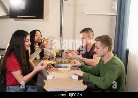 Company of four young friends sitting in kitchen and taking out pizza pieces Stock Photo